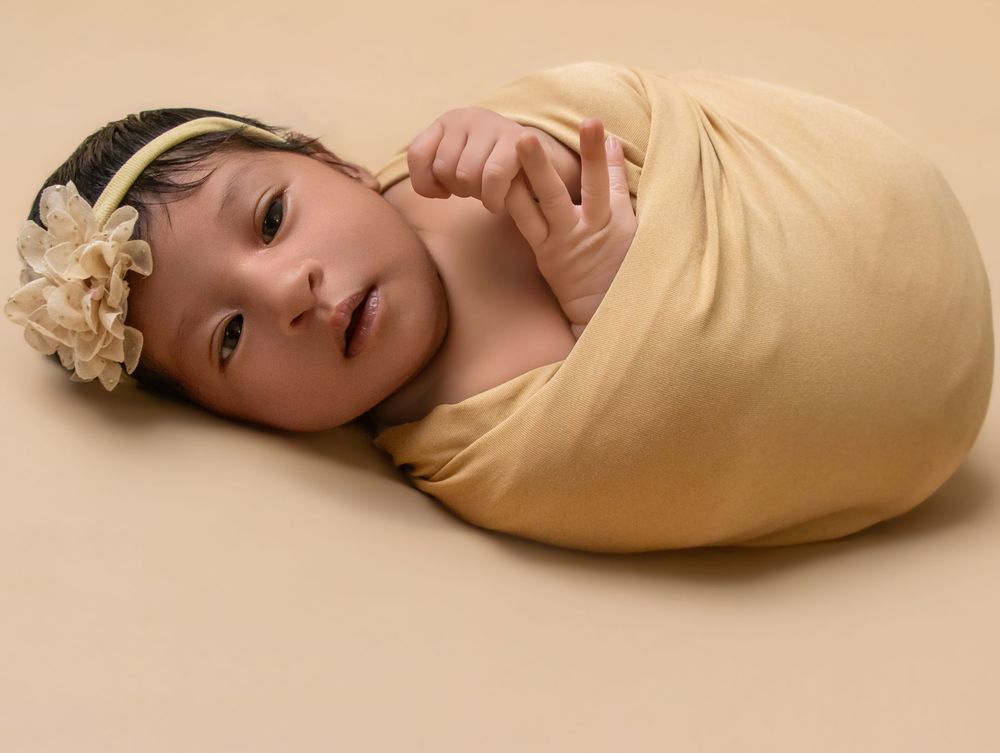 Newborn baby wrapped in a soft yellow swaddle blanket with a floral headband, lying on a beige background.