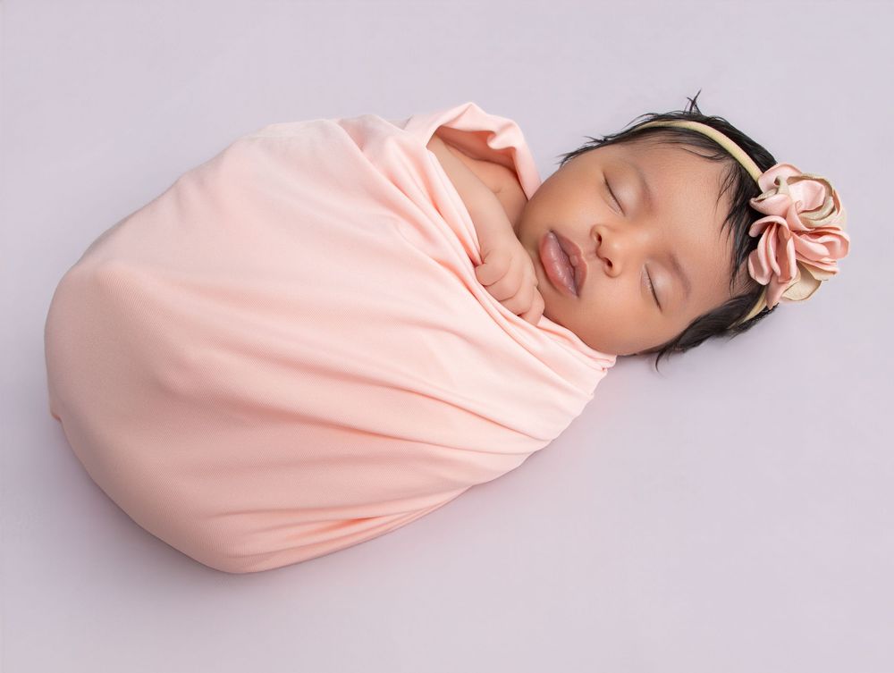 Newborn baby swaddled in a peach wrap, wearing a floral headband, sleeping peacefully on a soft gray background.
