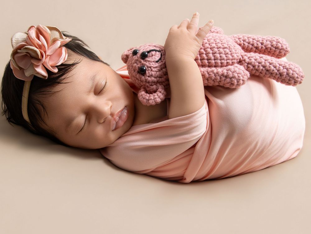 Sleeping newborn baby wrapped in pink cloth holding a crochet bunny toy, wearing a floral headband.