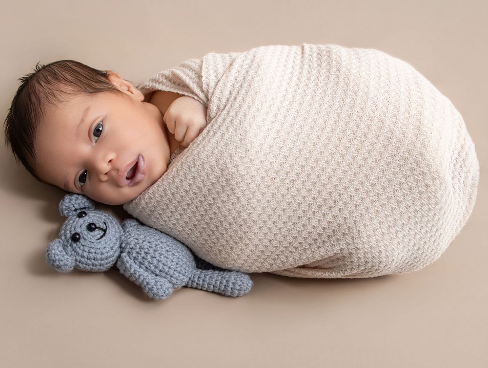 Newborn baby swaddled in cream wrap lying beside a blue crocheted teddy bear on a beige background.