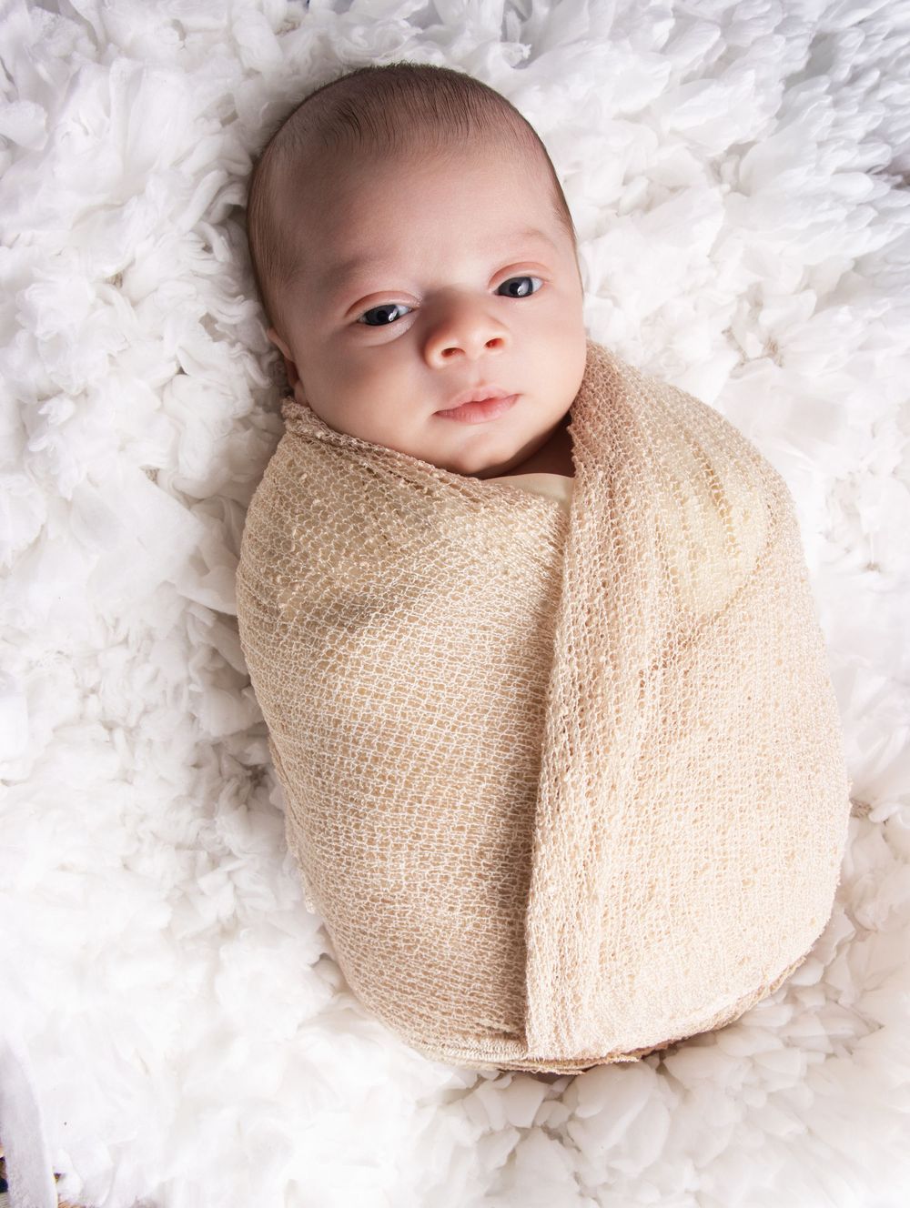 Newborn baby swaddled in a beige knit wrap, lying on a fluffy white blanket, looking up with bright eyes.