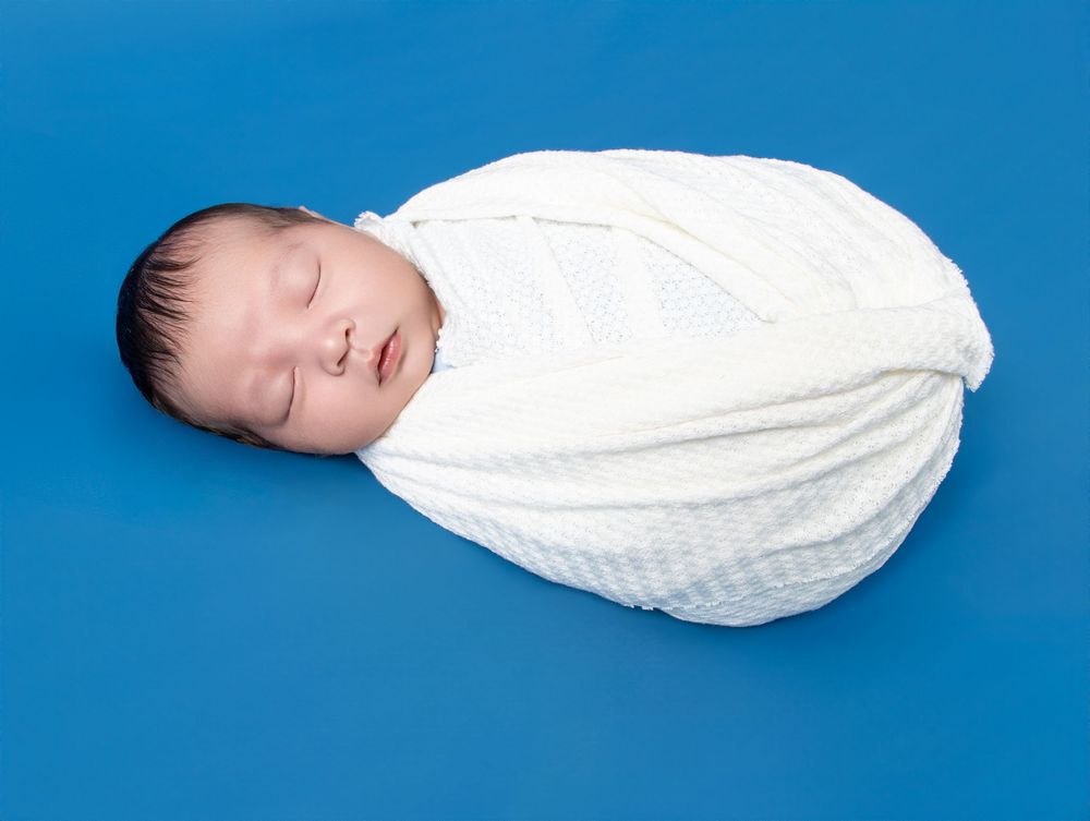 Peaceful newborn baby swaddled in white knit blanket sleeping on blue background.