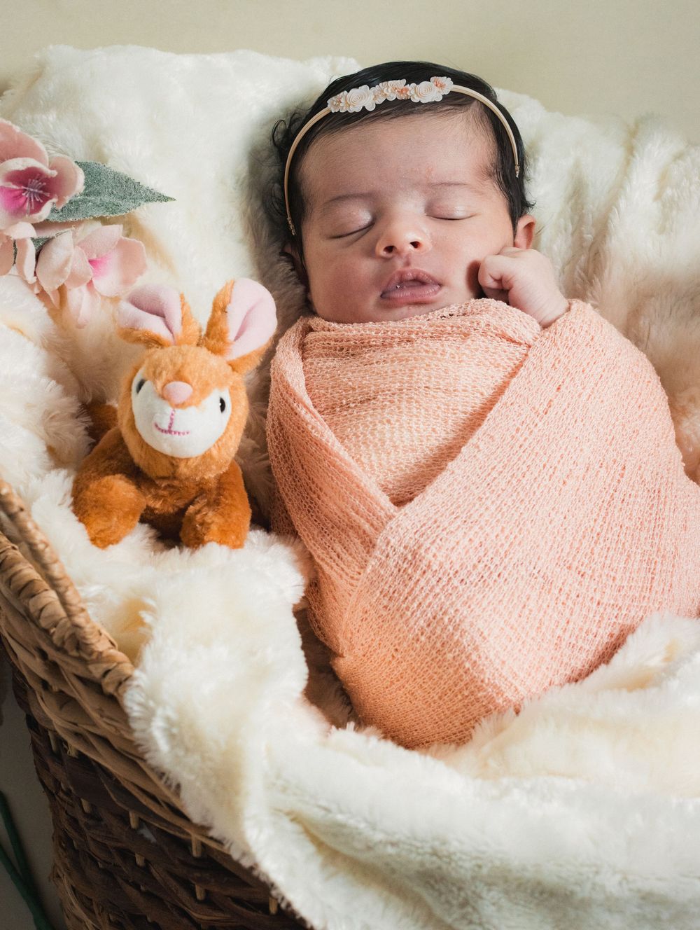Sleeping newborn baby swaddled in peach wrap with floral headband beside a stuffed deer toy in a wicker basket.