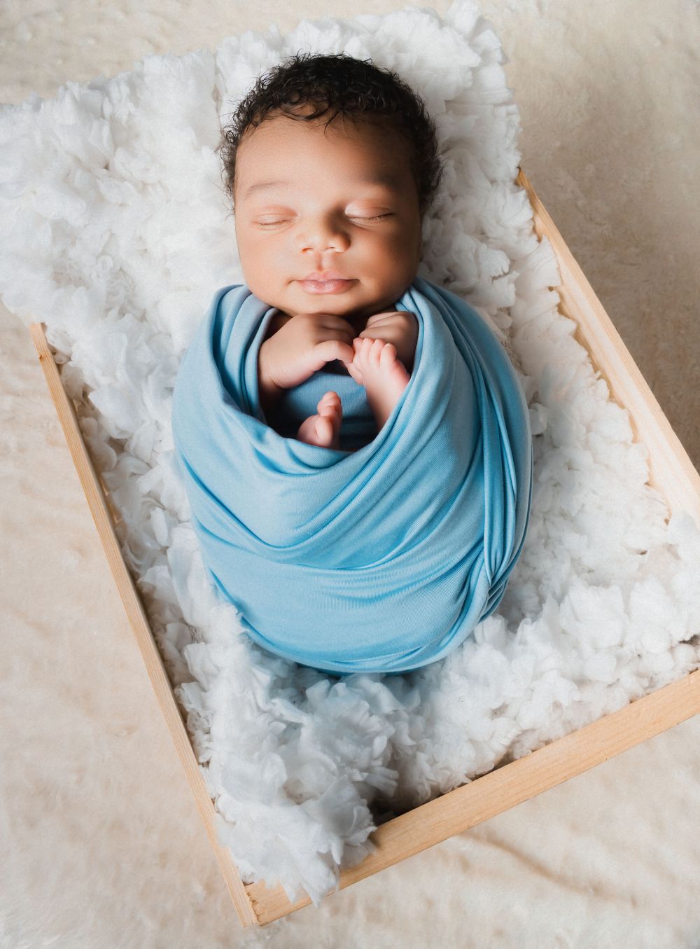 Peaceful newborn baby swaddled in a blue wrap, sleeping in a wooden crate lined with white fluffy material.