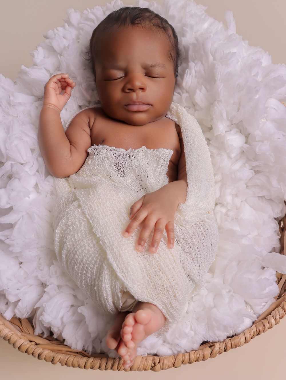 Sleeping newborn baby wrapped in white knit swaddle, nestled in a wicker basket with white floral bedding.