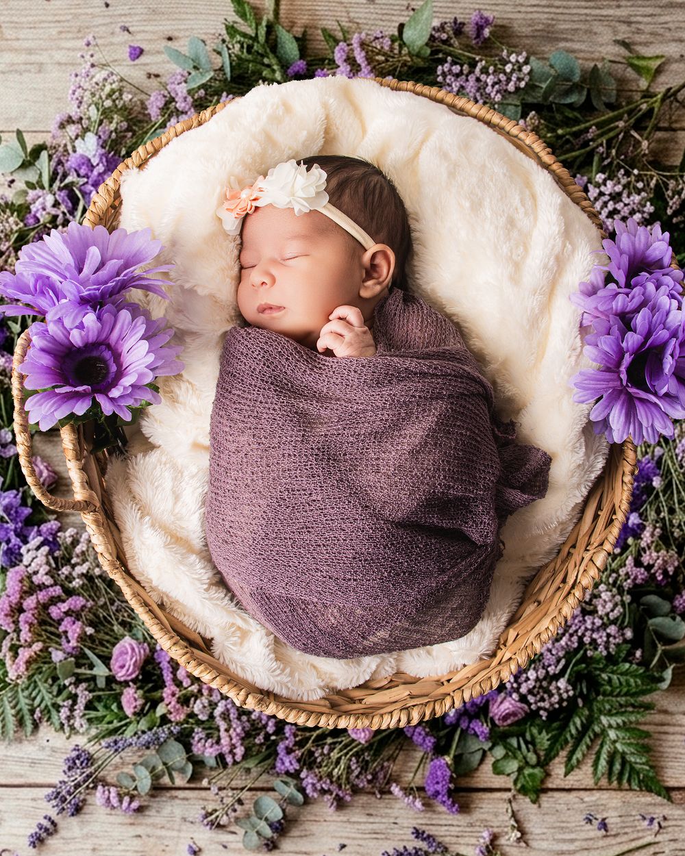 Sleeping newborn baby swaddled in purple wrap nestled in wicker basket surrounded by lavender and purple flowers.