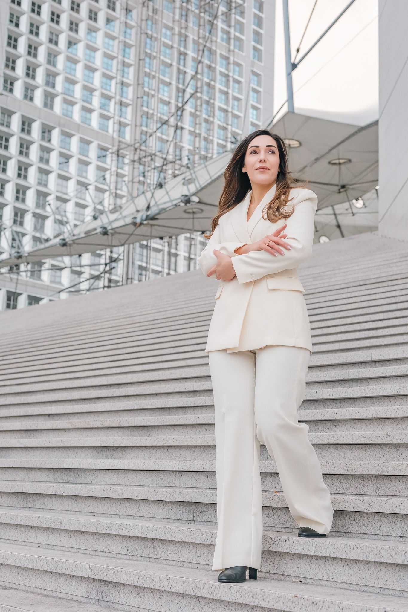 Confident businesswoman in white suit stands with arms crossed on steps outside a modern office building.