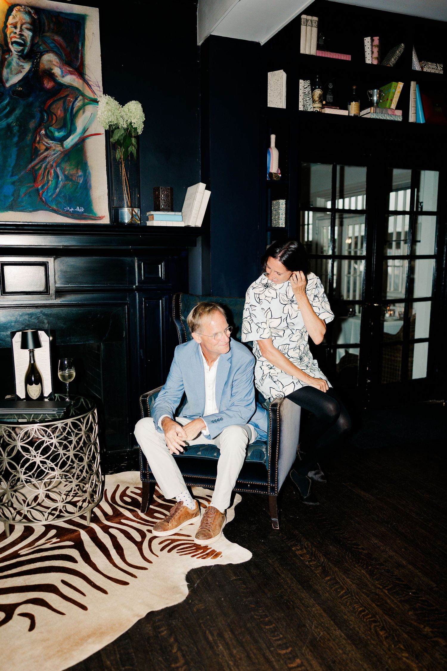 Two people relaxing in a dark, elegantly decorated living room with zebra rug, artwork, and champagne.