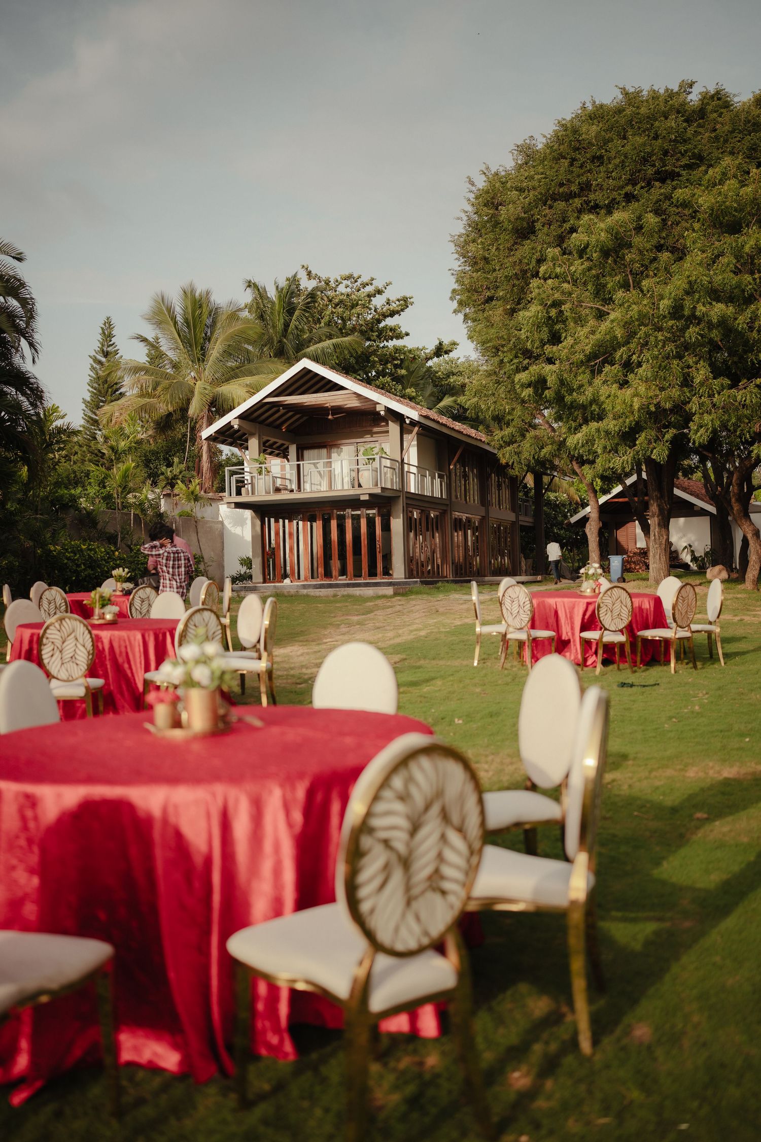 Outdoor wedding reception on green lawn with red-draped round tables, elegant white chairs, and tropical wooden venue backdrop.