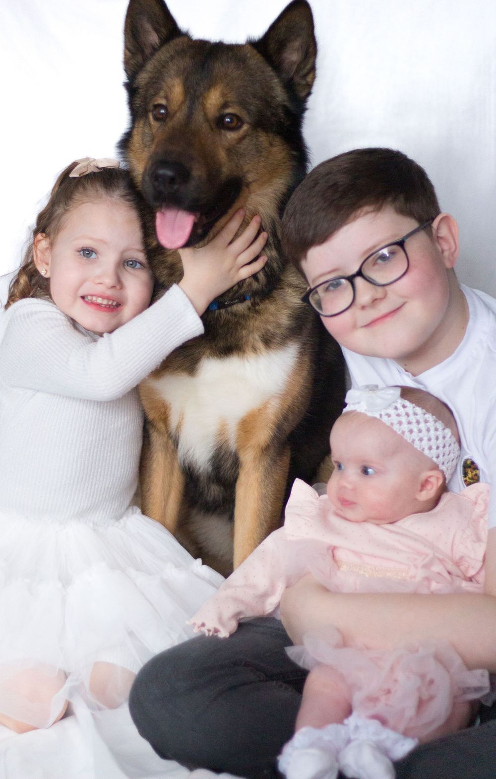 Two children and a baby pose with a German Shepherd dog against a white background.