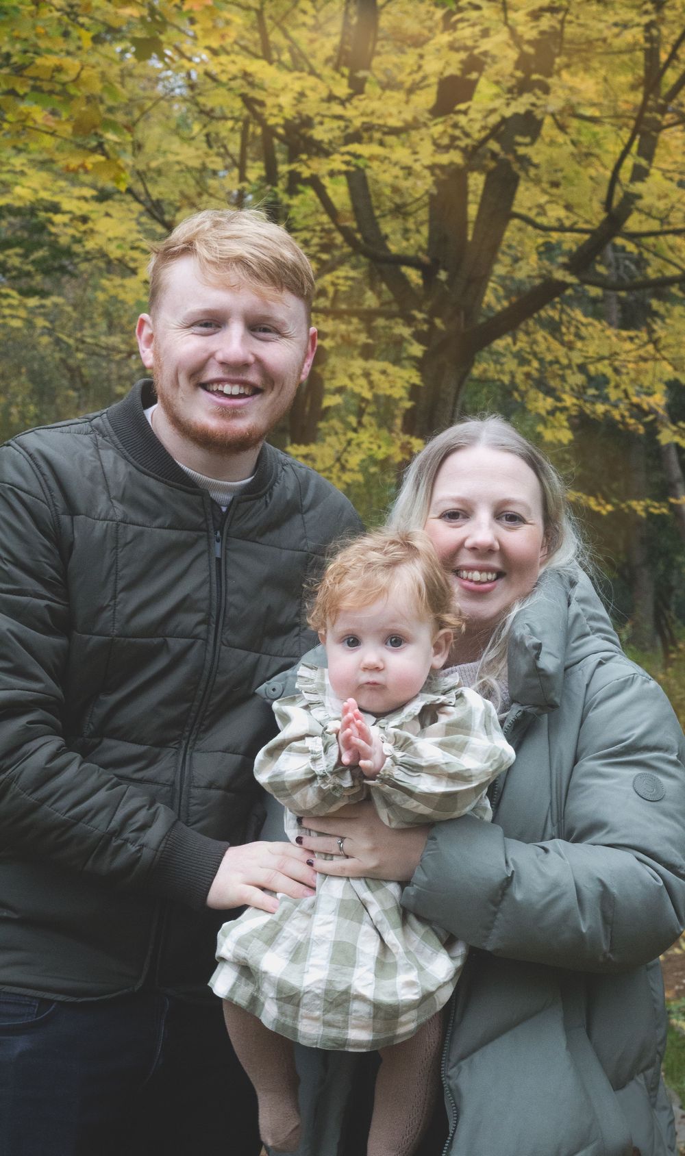 A smiling couple holds their red-haired toddler daughter outdoors among autumn yellow trees.