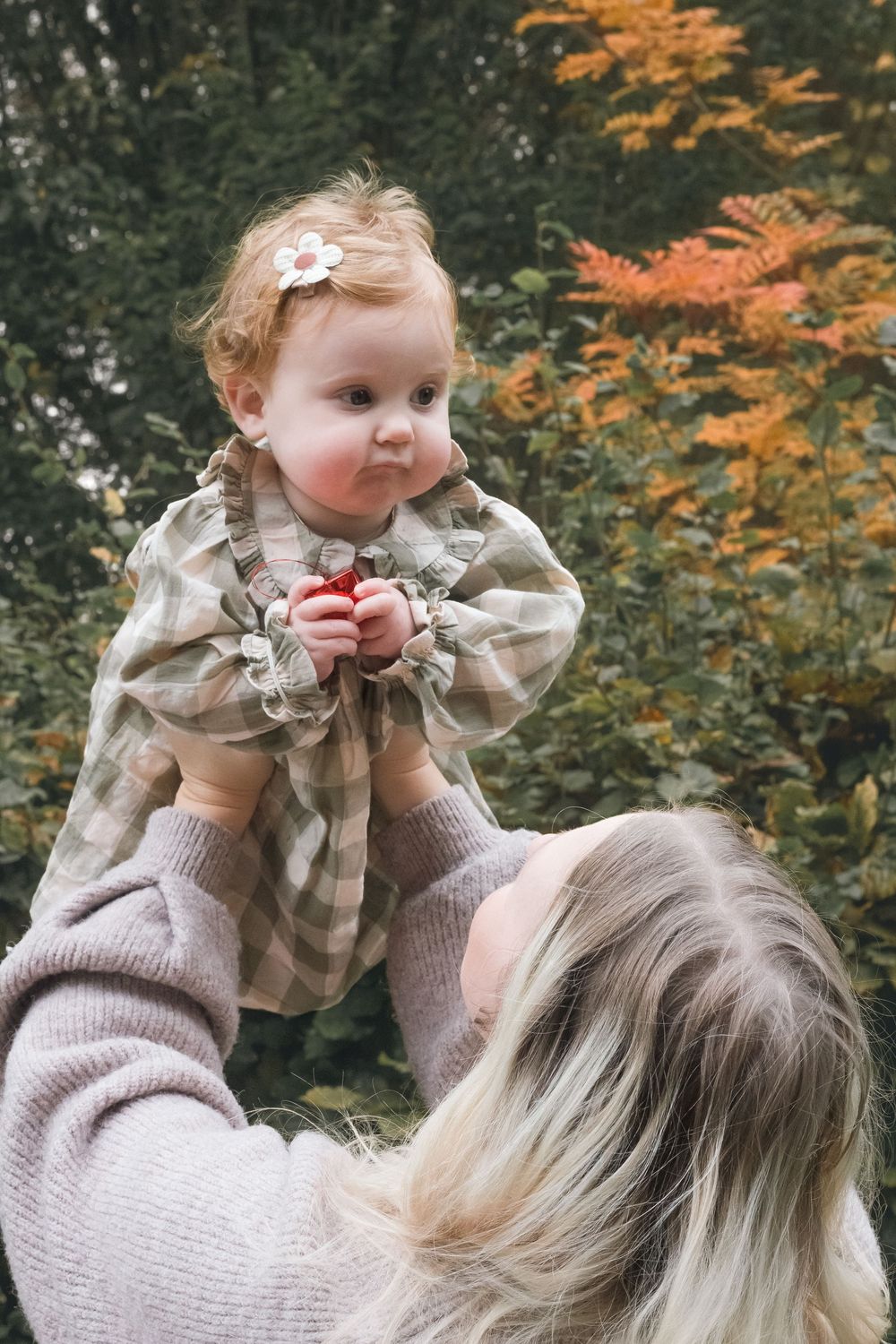 A blonde mother lifts her baby girl in a plaid dress with a daisy hair clip outdoors.