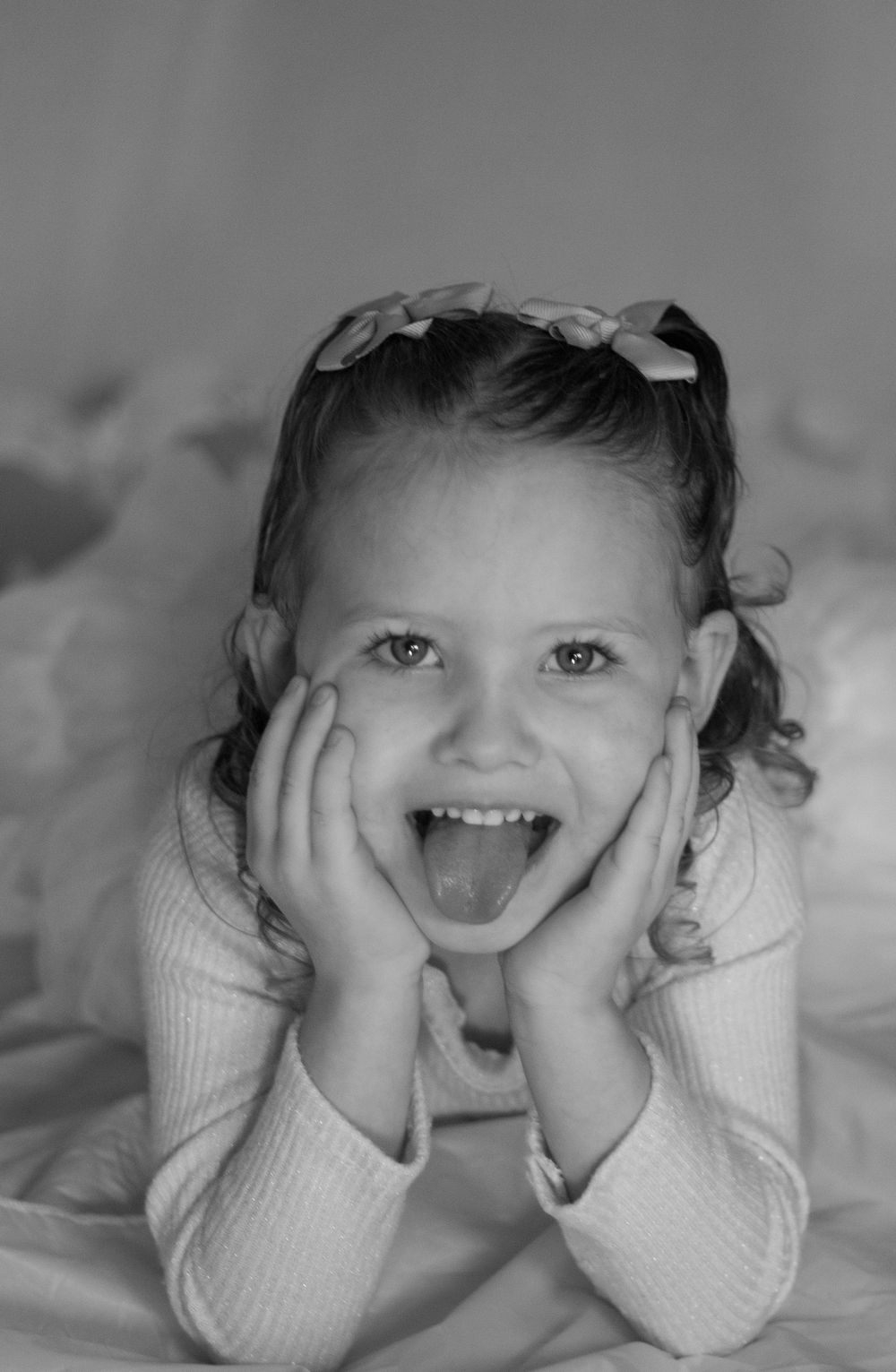 A young girl with pigtails makes a funny face, holding her cheeks with both hands while sticking out her tongue.