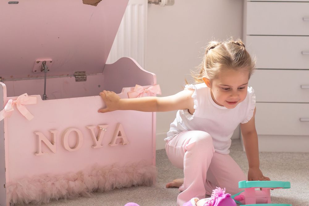 Young girl playing beside a pink personalized toy chest labeled NOVA in a soft pink girls bedroom.