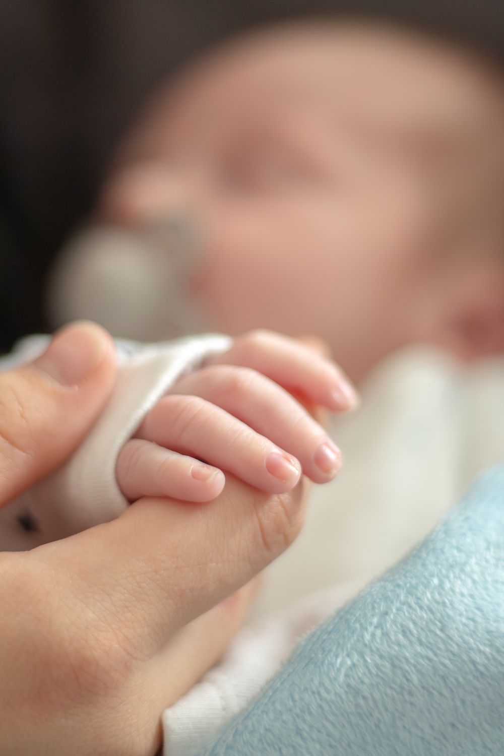 A newborn baby grasps an adult finger while resting, showcasing the tiny delicate hand of an infant.
