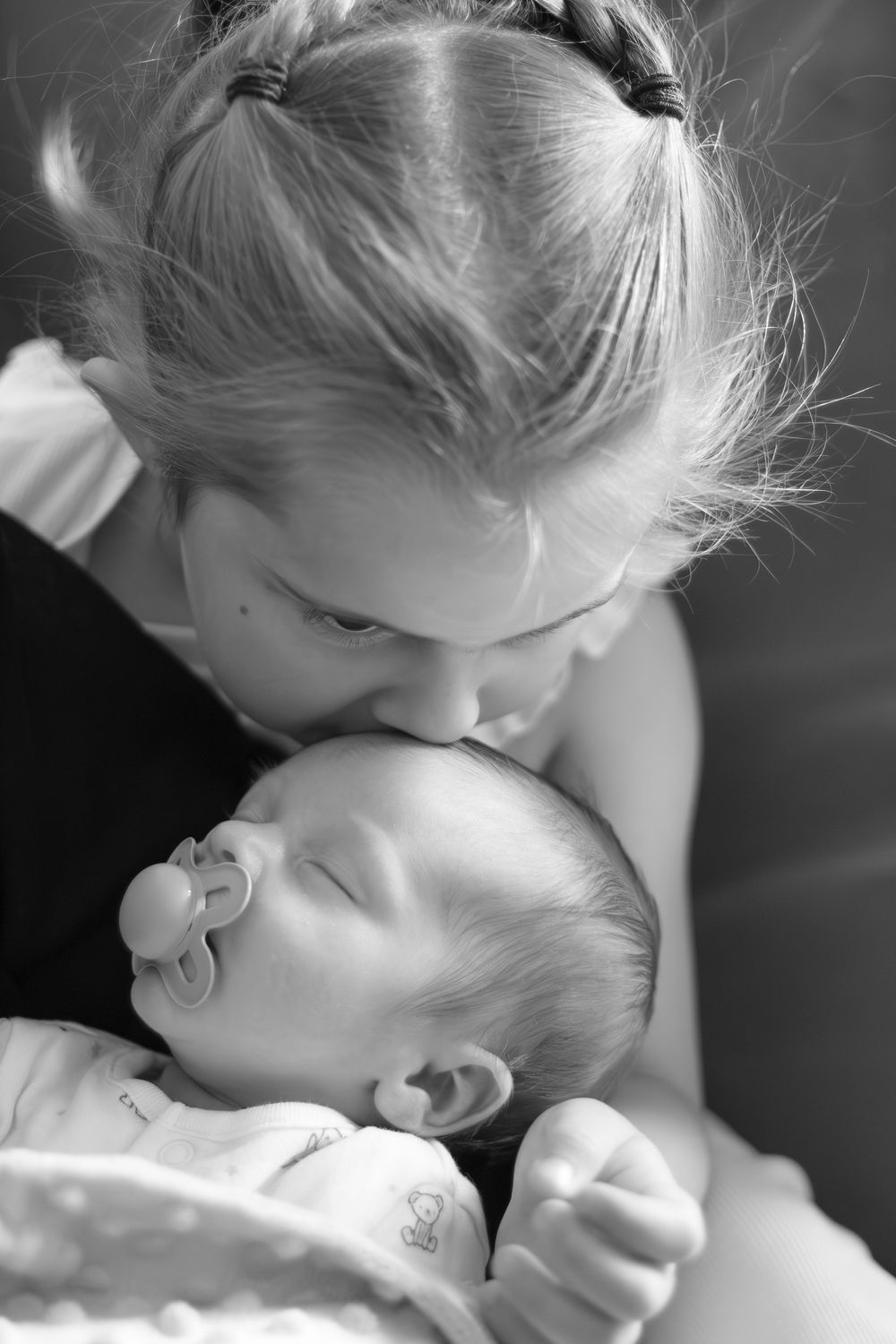 Black and white photo of a young girl gently kissing a sleeping newborn baby with a pacifier.