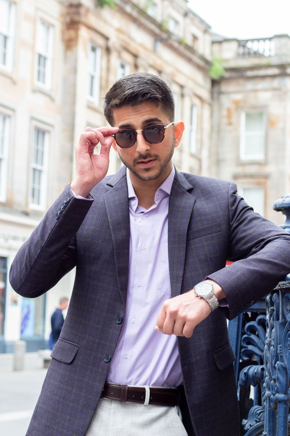 Stylish young man in grey suit adjusting sunglasses on urban street with historic buildings.