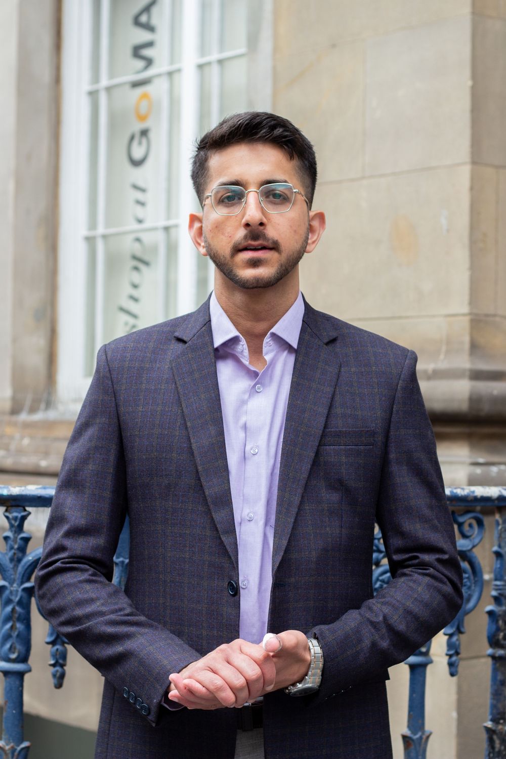 Confident young man in grey checked suit and glasses standing by ornate blue railings outdoors.