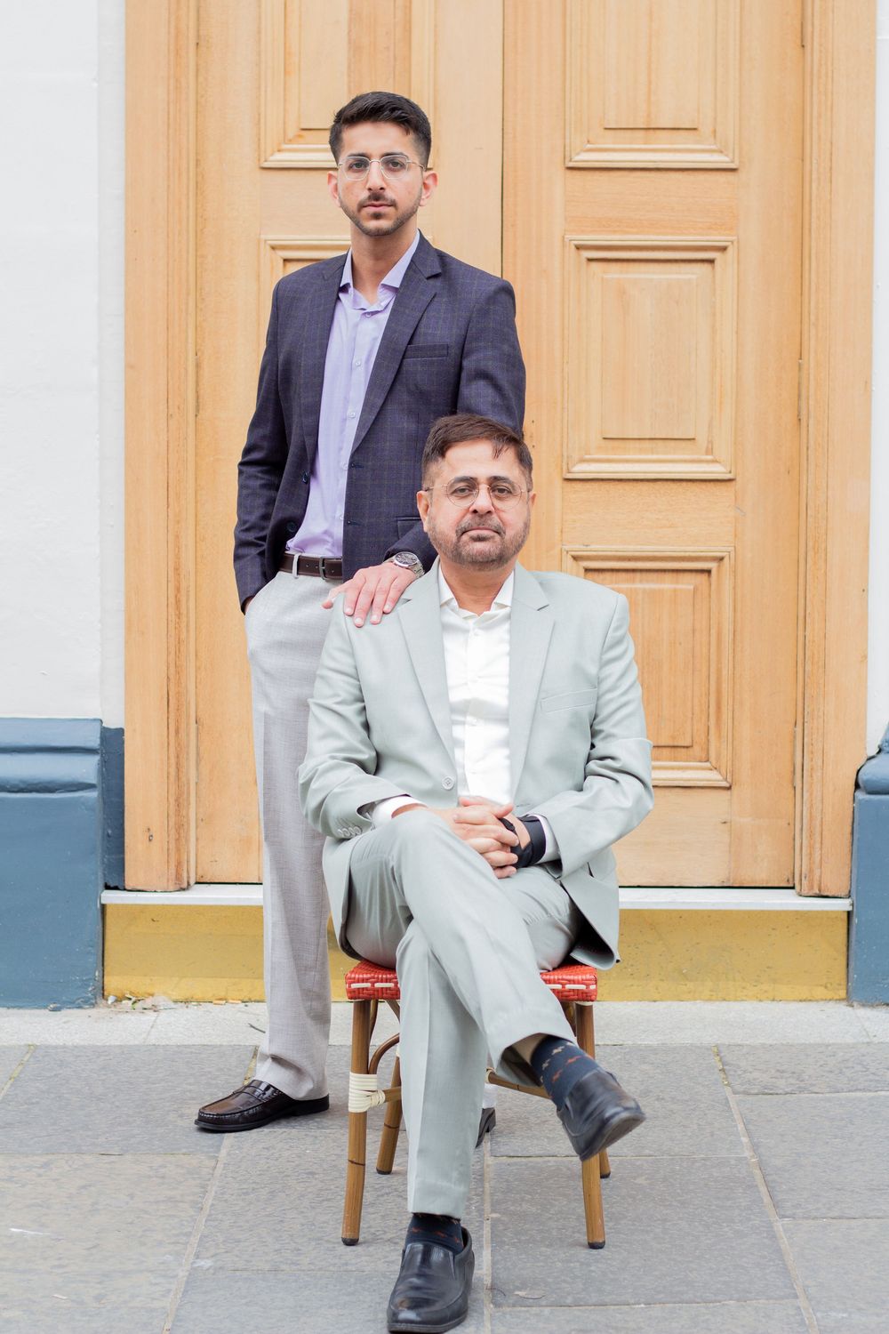 Two men in suits pose formally, one seated on a chair and one standing, in front of a wooden door.
