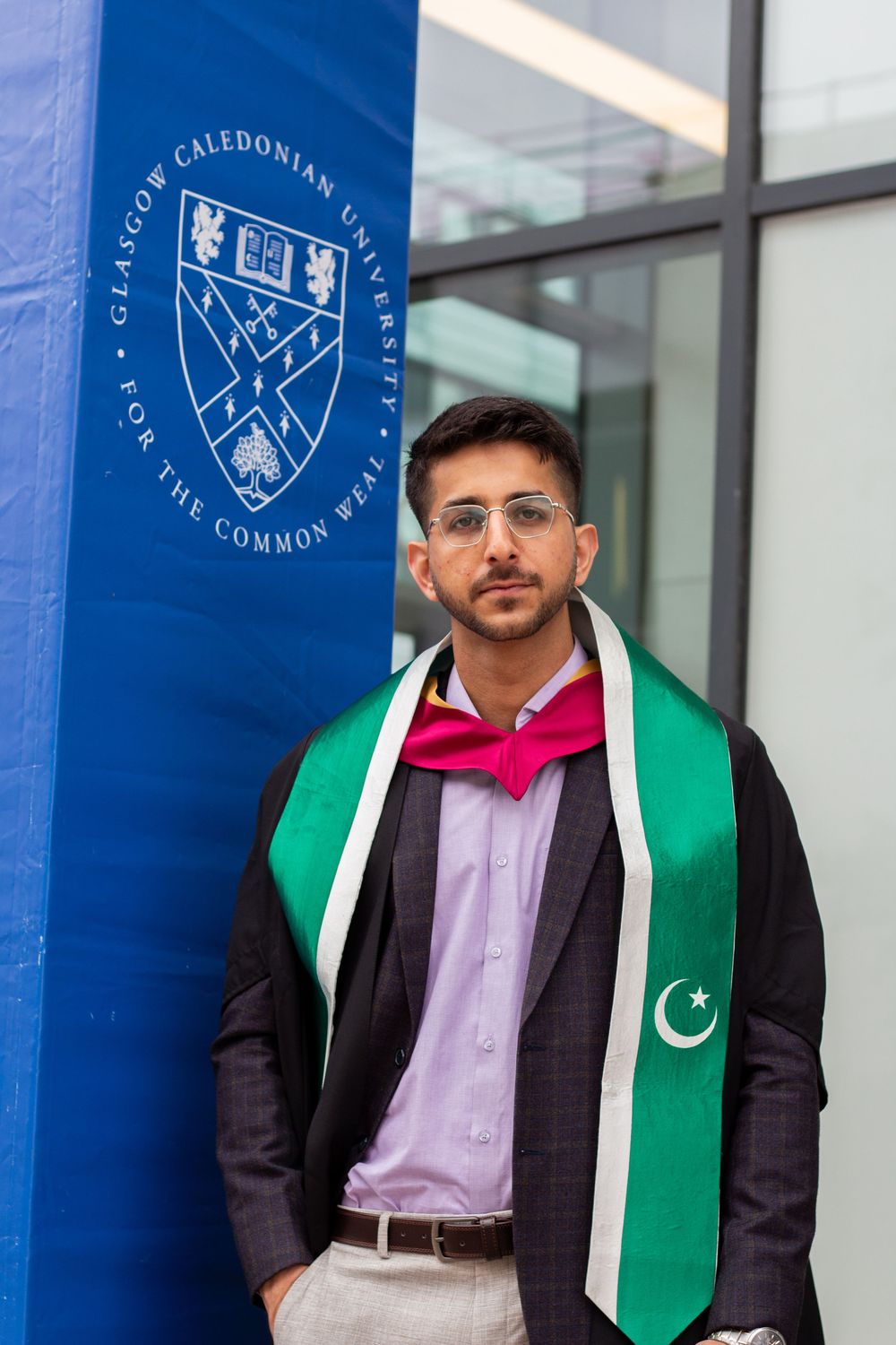 Young man in graduation attire with Pakistani flag sash poses by Glasgow Caledonian University sign.