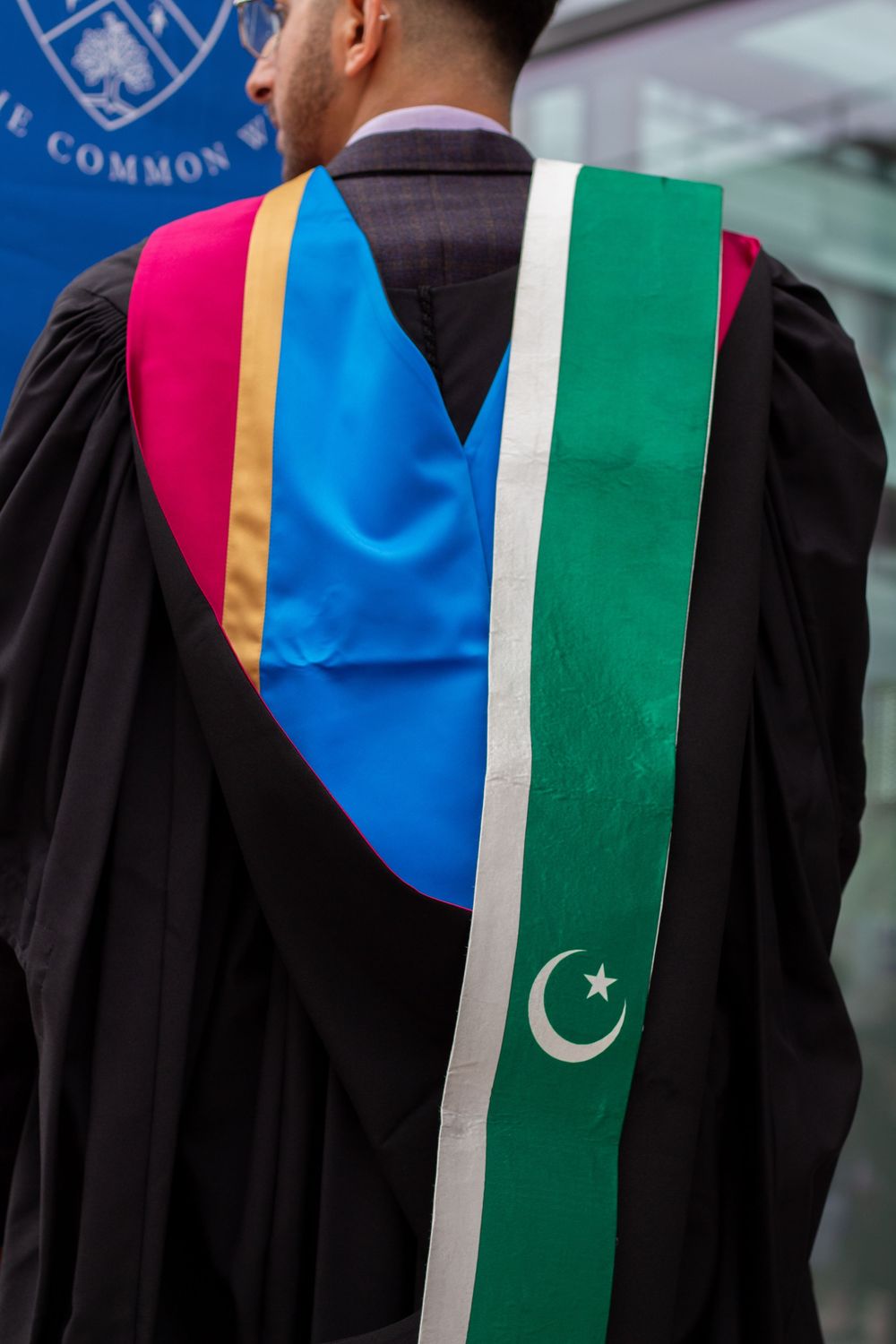 Graduate in black academic gown with colorful hood and Pakistani flag sash at Glasgow Caledonian University ceremony.