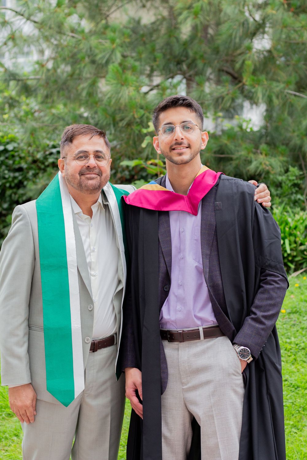 Father and son smiling together outdoors, son wearing graduation gown and pink hood.