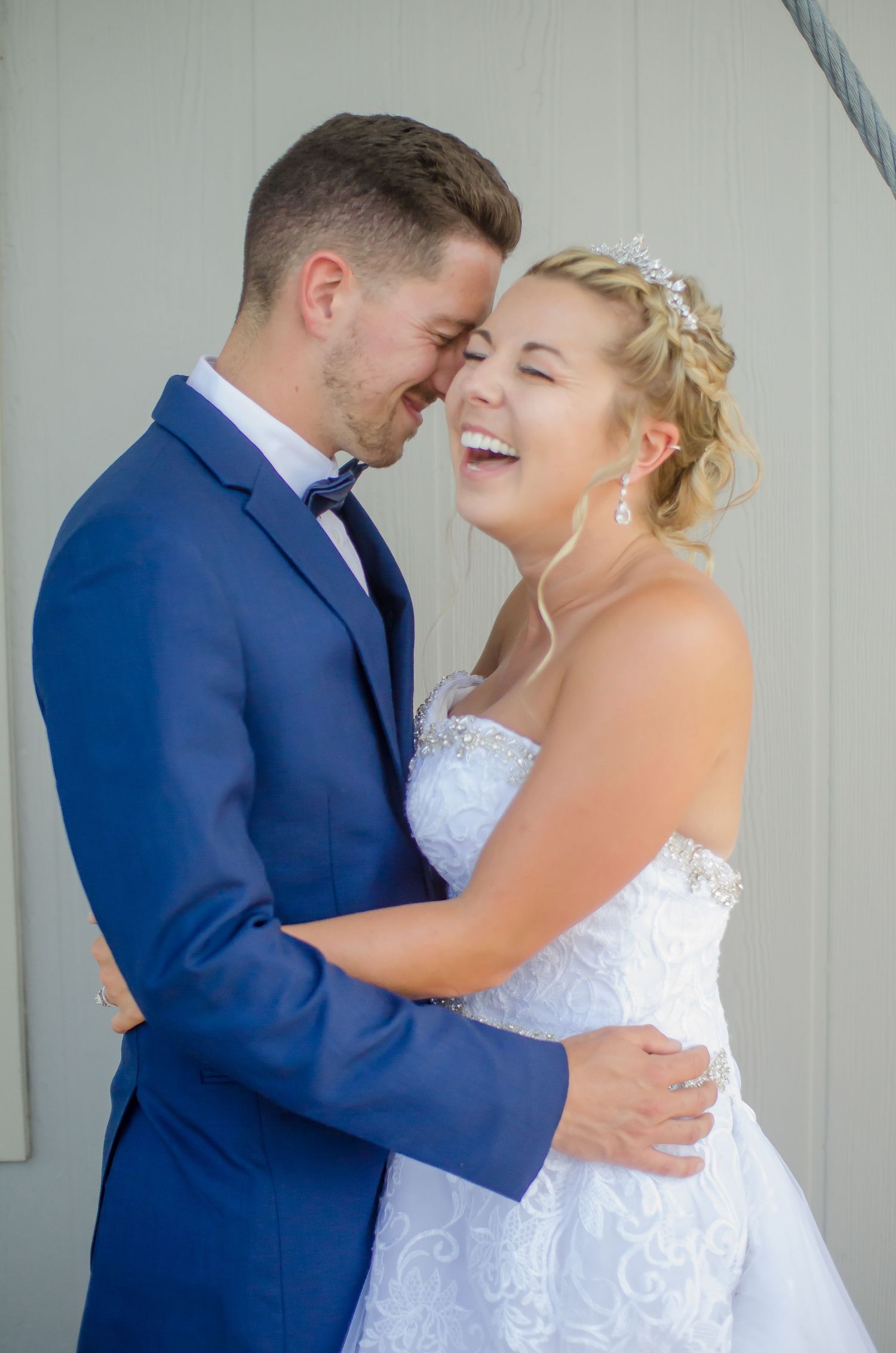 Joyful bride in white lace gown and groom in blue suit share a laughing embrace on their wedding day.