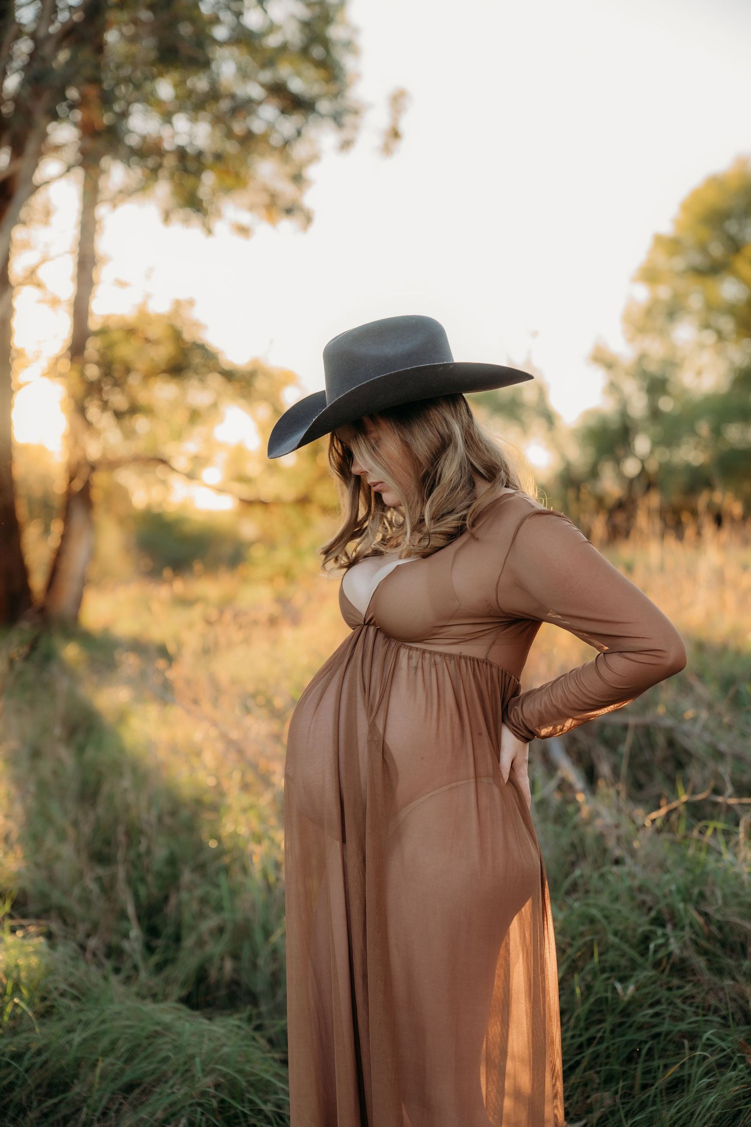 Pregnant woman in brown maxi dress and wide-brim hat poses outdoors at golden hour in a natural field setting.