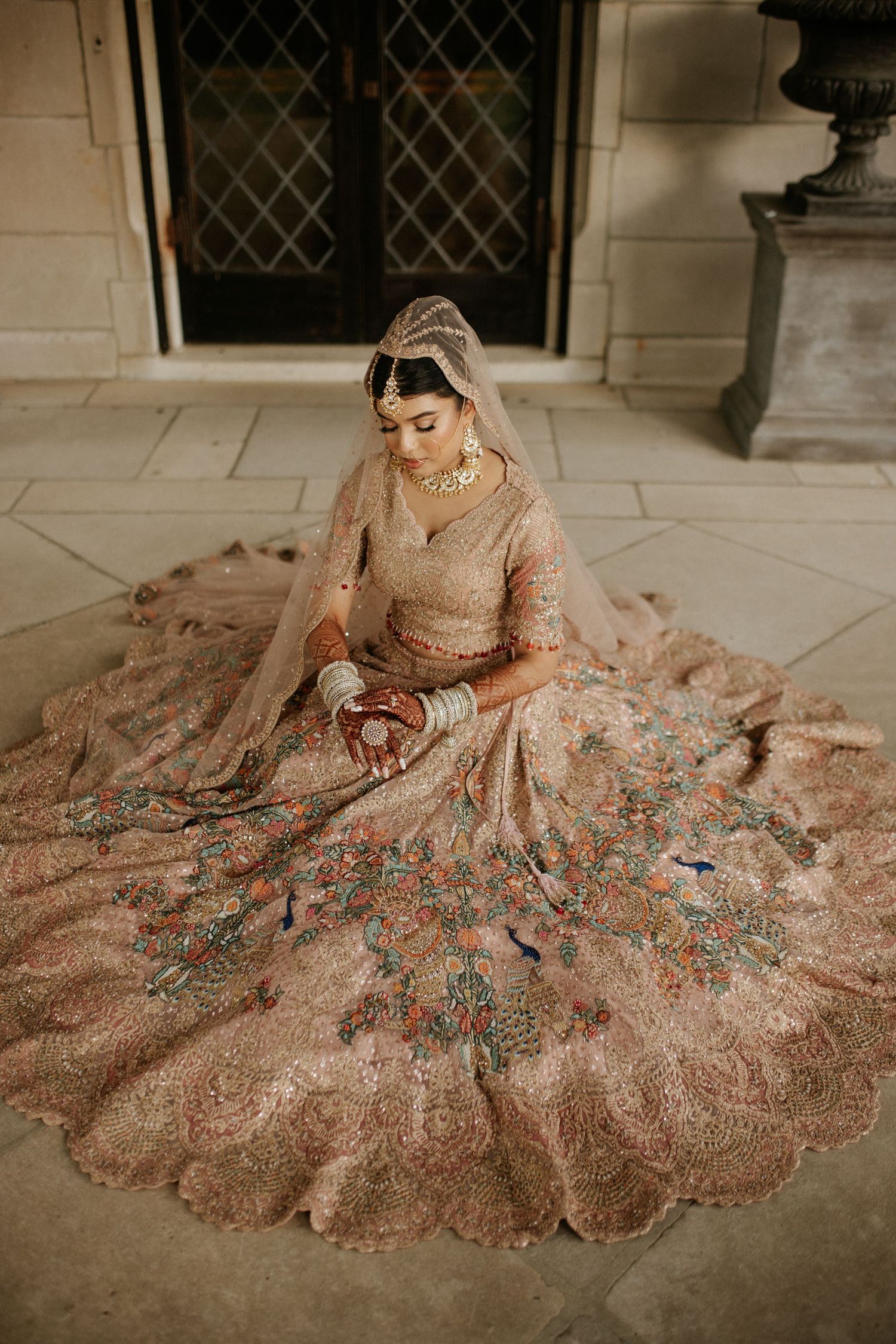 Bride in ornate dusty rose lehenga with colorful floral embroidery sits gracefully on floor wearing gold jewelry and dupatta.