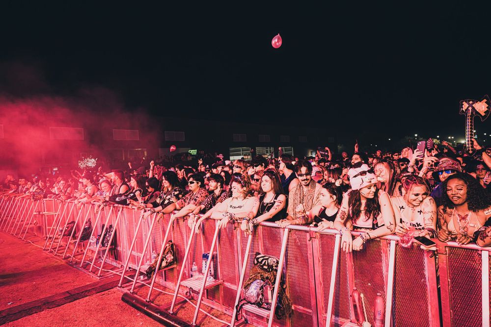 Large crowd of concert fans pressed against metal barriers under red stage lighting at an outdoor night festival.