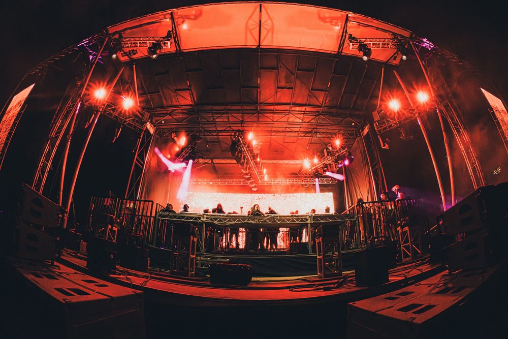 Wide-angle fisheye view of a concert stage with red and purple lighting rigs above a DJ booth at a live music event.