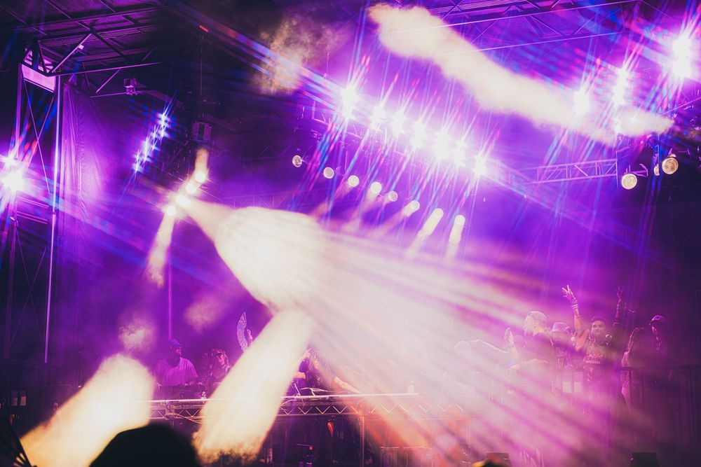 Concert stage with vibrant purple laser lights and smoke effects illuminating a performer before a live crowd.