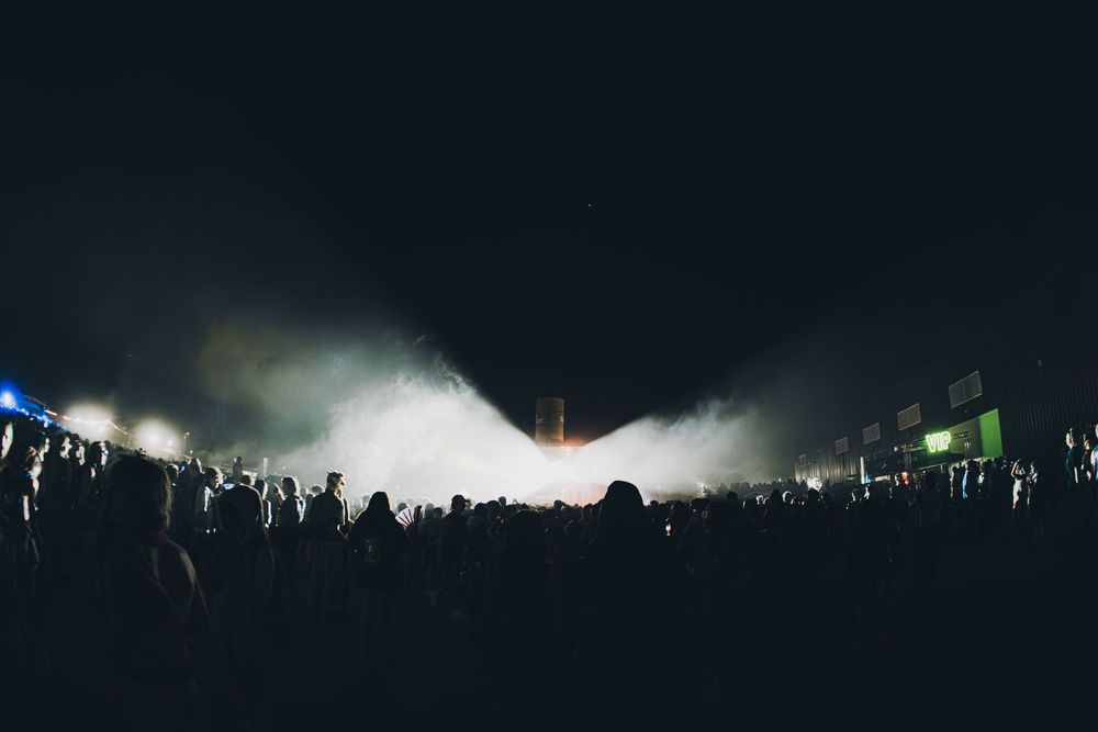 Large crowd at an outdoor night music festival with dramatic fog and bright stage lights illuminating the dark sky.