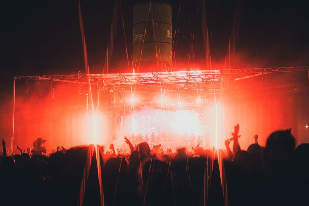 Concert crowd silhouetted against vibrant red stage lights with laser beams at a live music festival.