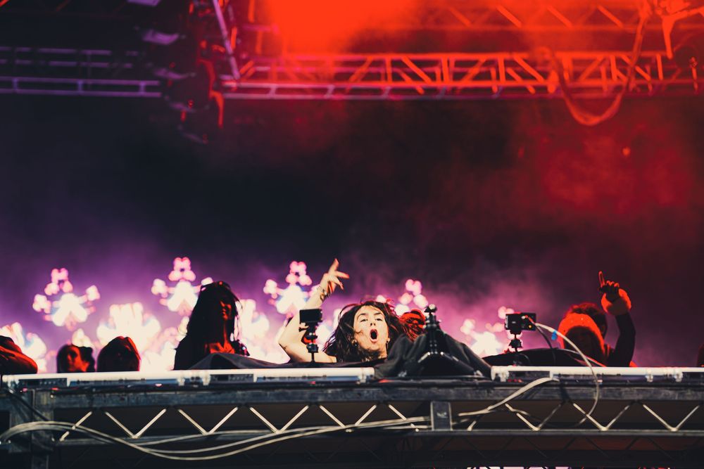 DJ performing energetically on stage with vibrant purple and pink lights illuminating the crowd at a live music festival.