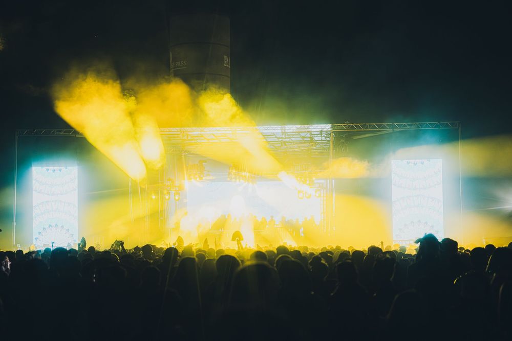 Concert crowd silhouetted against vibrant yellow smoke and bright stage lights at an outdoor music festival.