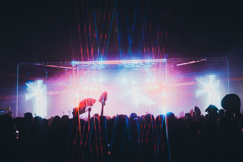 Concert crowd silhouetted against vibrant pink and blue laser light show on a dark stage.