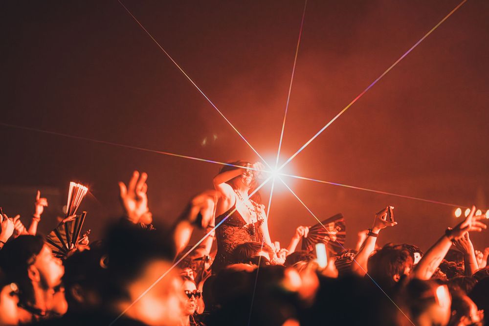 Concert crowd under bright laser lights with red-toned atmosphere at a live music festival event.