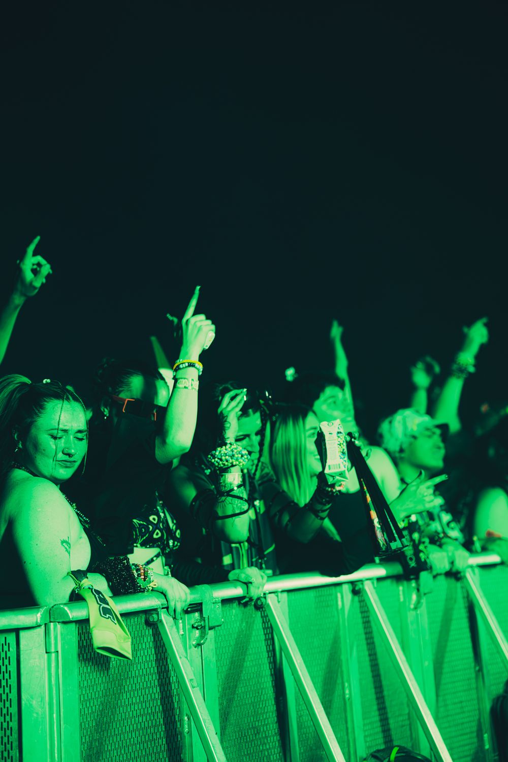 Excited concert crowd bathed in green light, fans raising hands and cheering behind metal barriers at a night music festival.