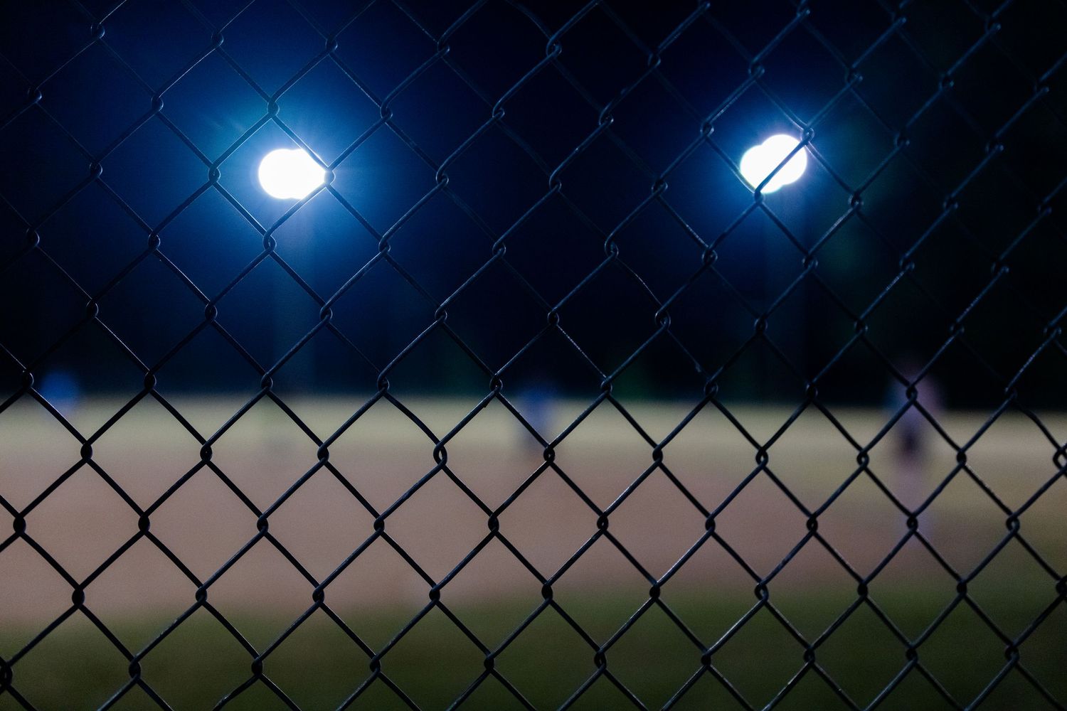 Chain-link fence at night with bright stadium lights illuminating a sports field in the background.