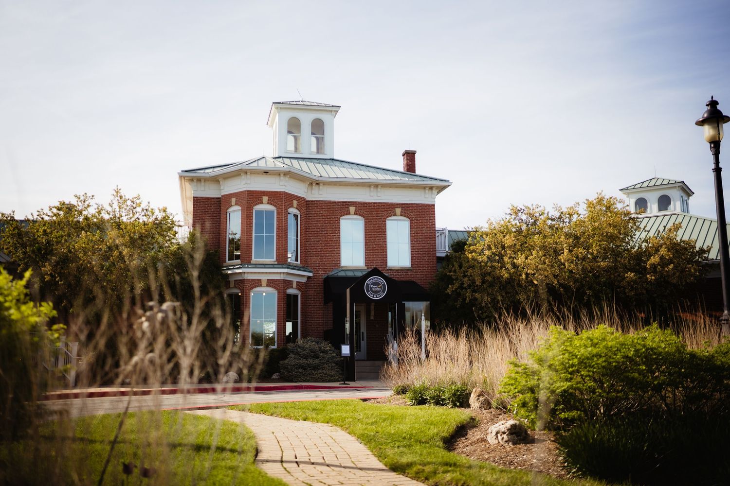 Historic red brick building with white cupola surrounded by lush greenery and brick pathway on a sunny day.