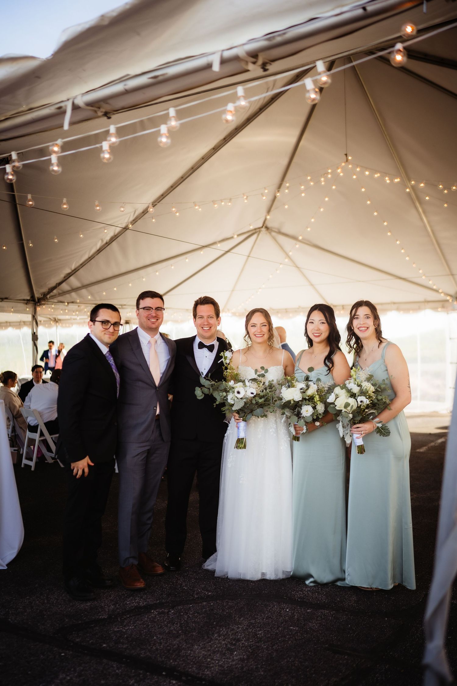 Wedding party posing under a tent with string lights, bride in white gown with bouquet, bridesmaids in sage green dresses.