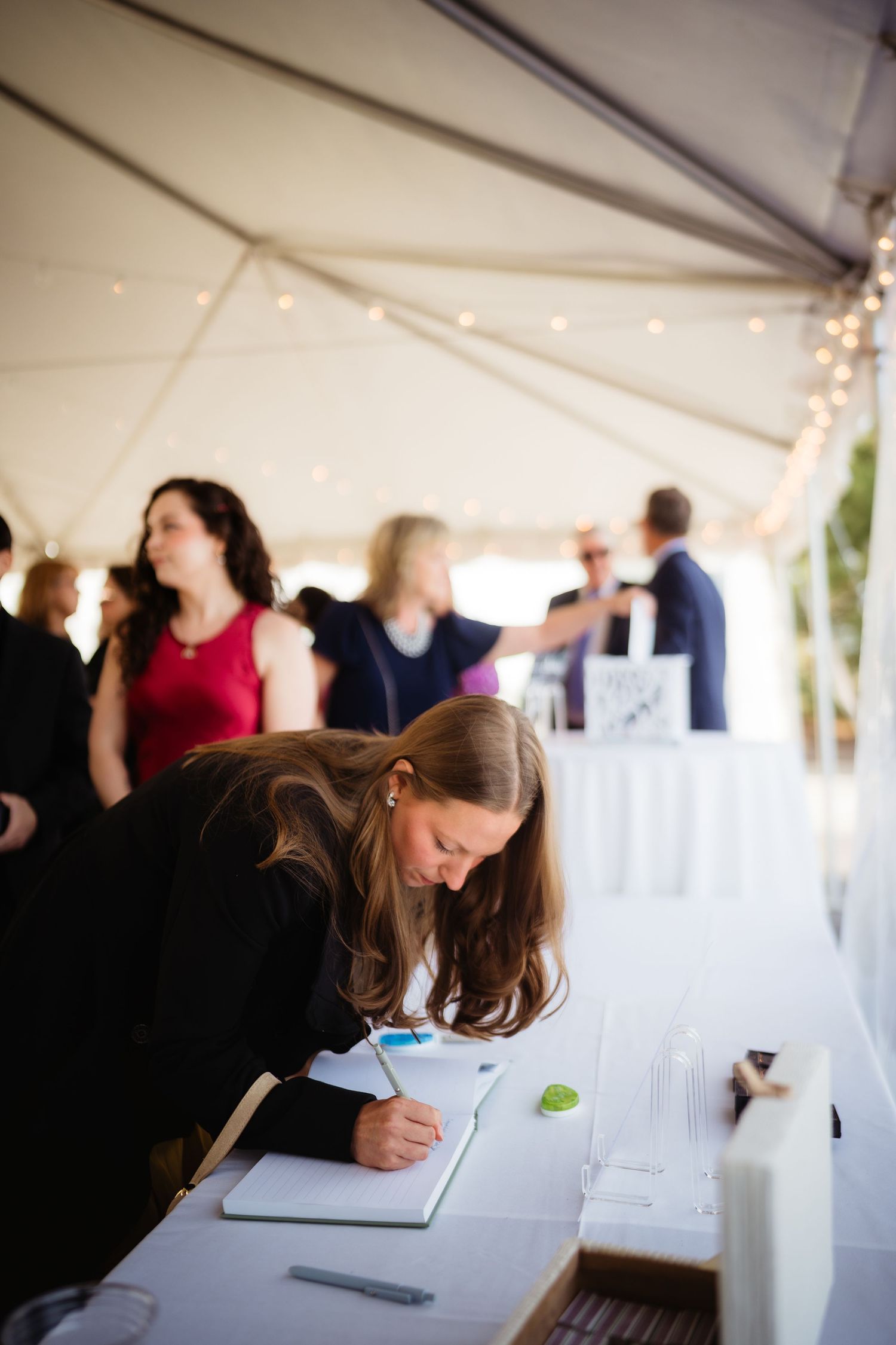 Woman signing guest book at elegant outdoor wedding reception under white tent with string lights.