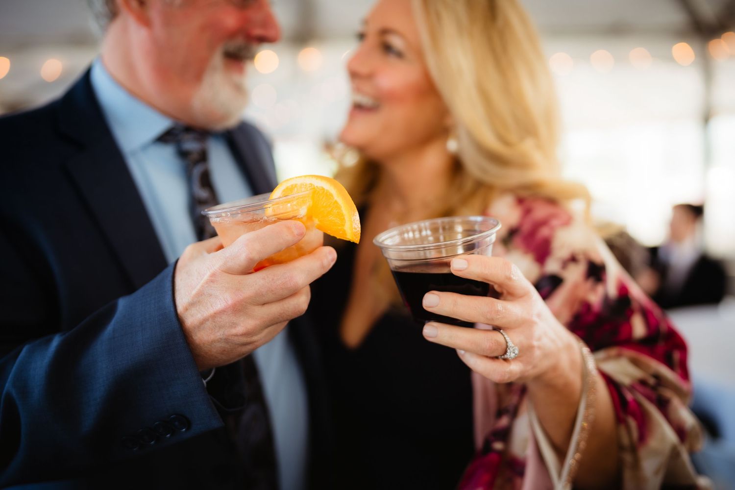 Couple smiling and toasting with cocktails at an elegant outdoor event, man holding citrus drink, woman with dark beverage.