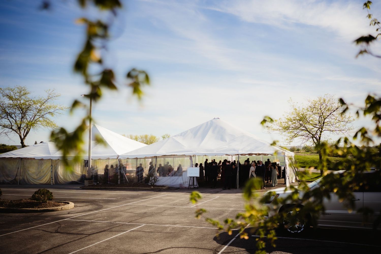 Elegant white pole tent wedding reception setup in a parking lot under a blue sky with scattered clouds.