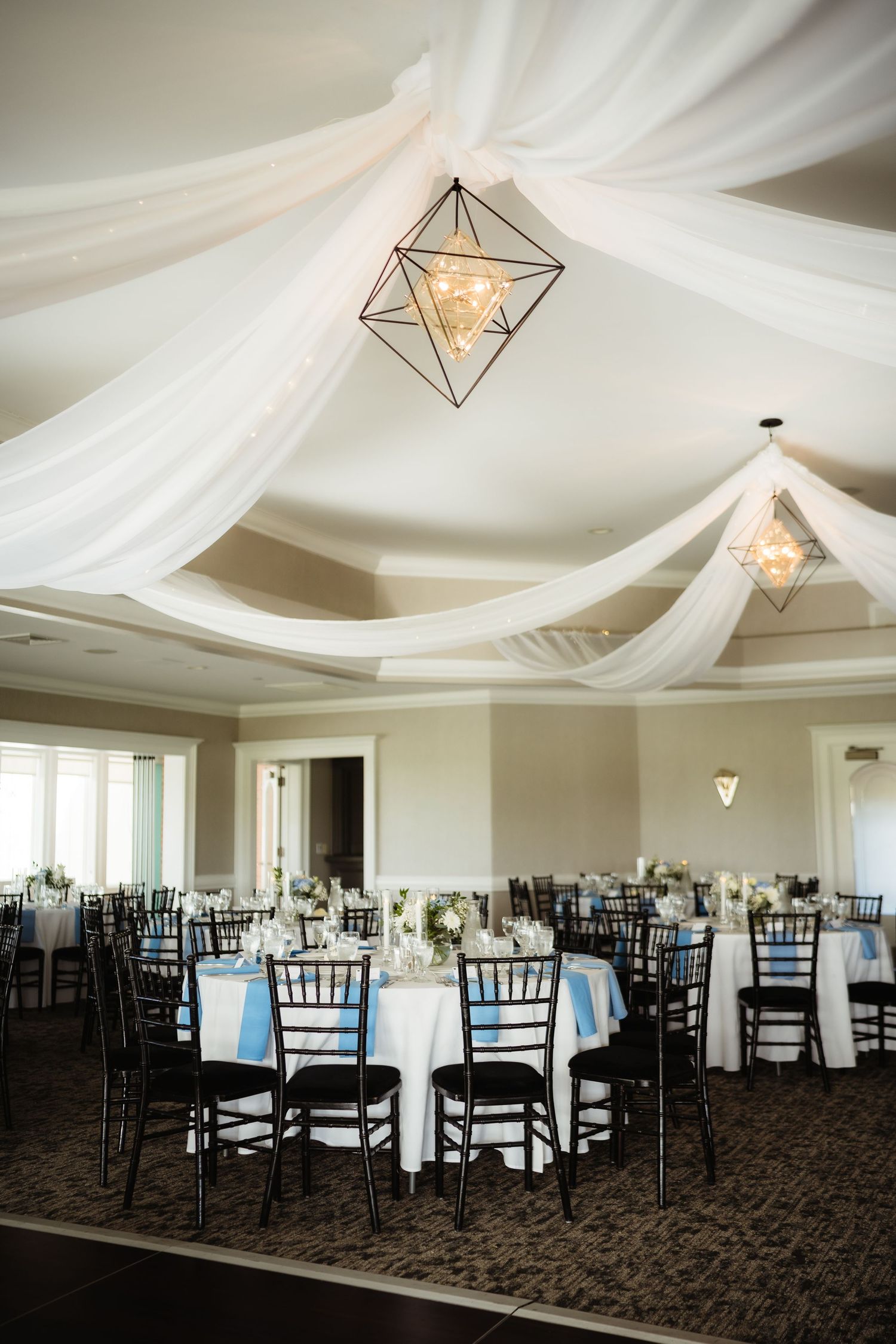 Elegant wedding reception hall with white draped ceiling, geometric pendant lights, and round tables with blue accents.