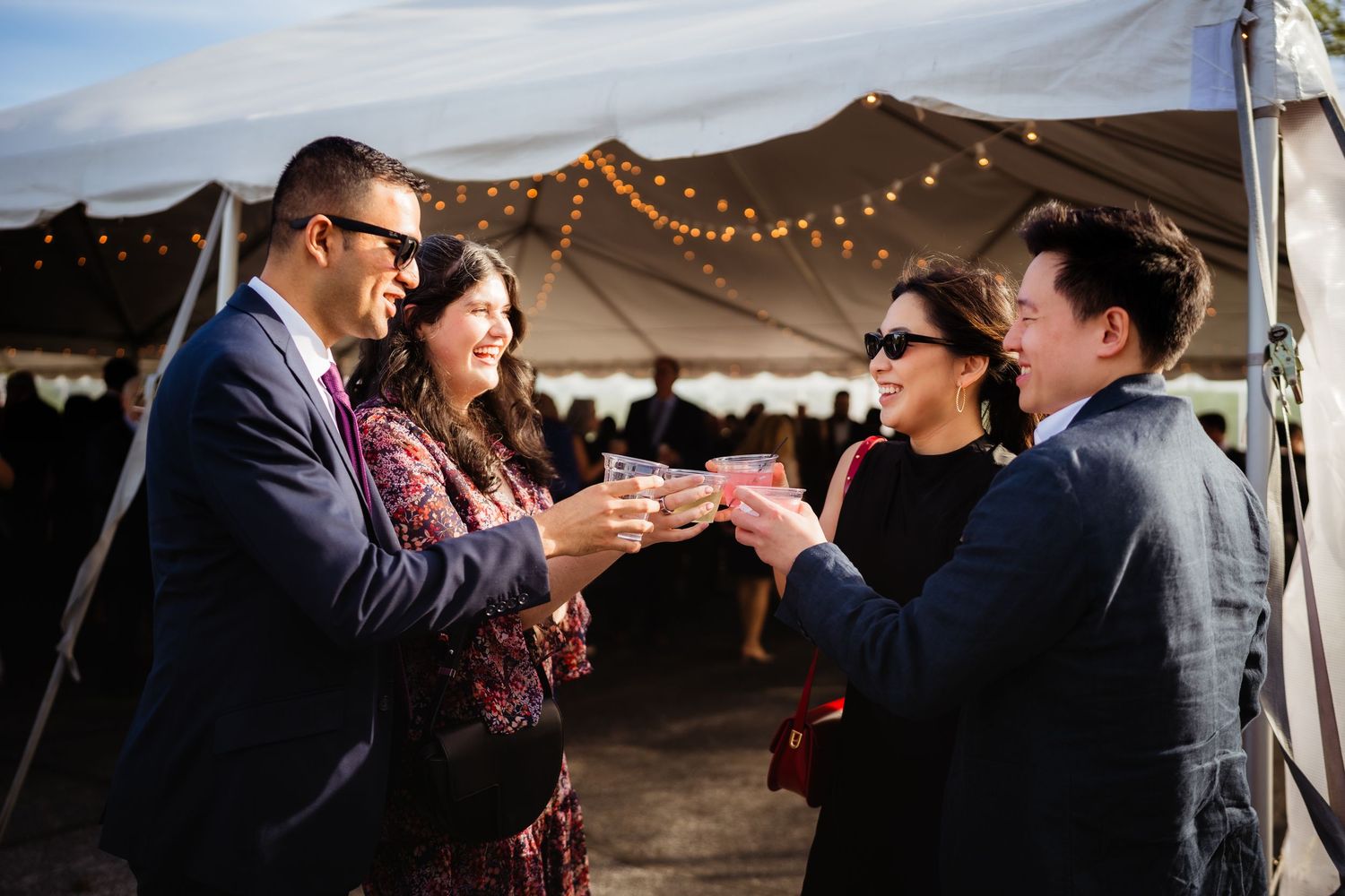 Four guests toasting with drinks under a white tent with string lights at an outdoor wedding reception.