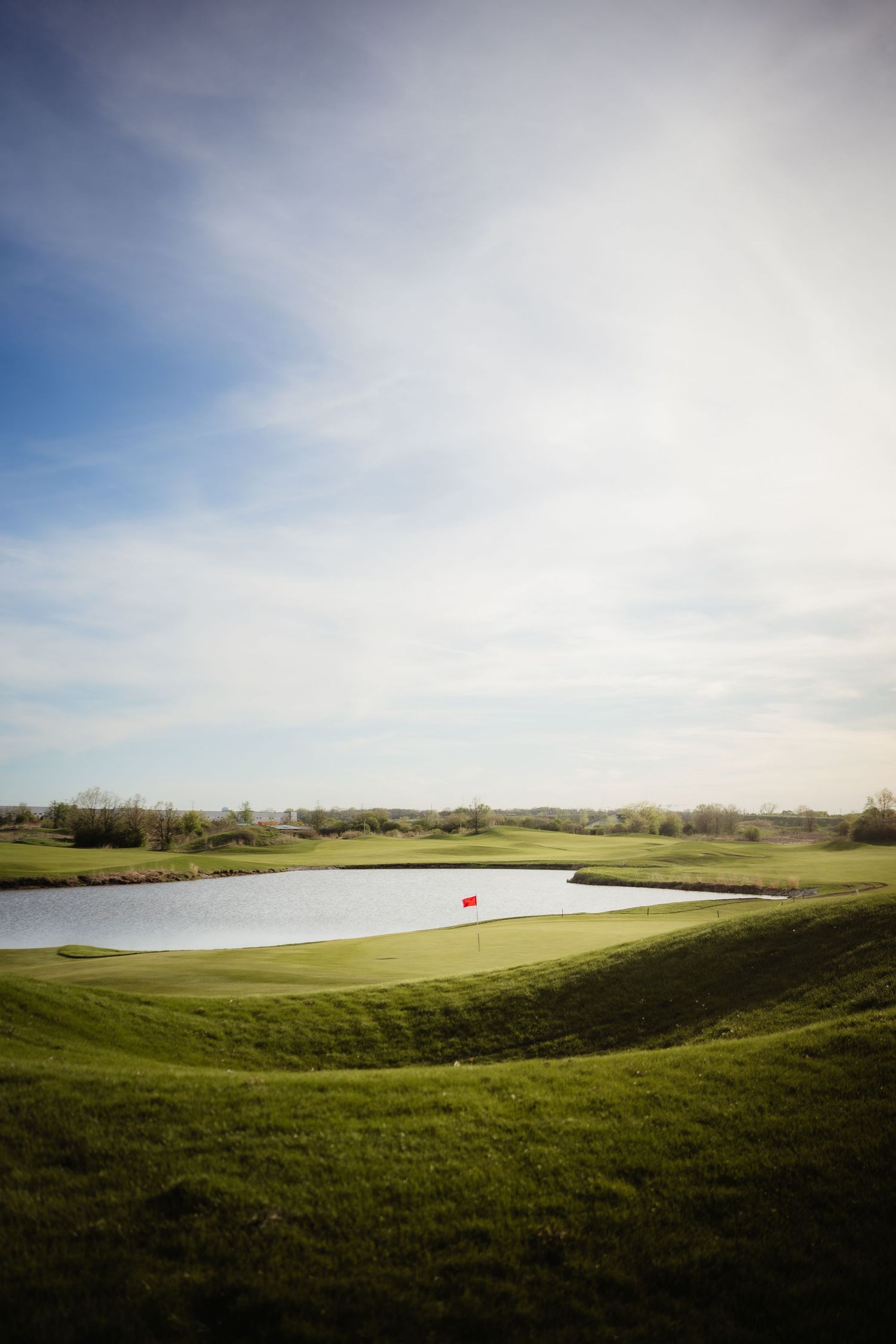 Scenic golf course with lush green fairways, a serene water hazard, and a red flag pin under a partly cloudy sky.