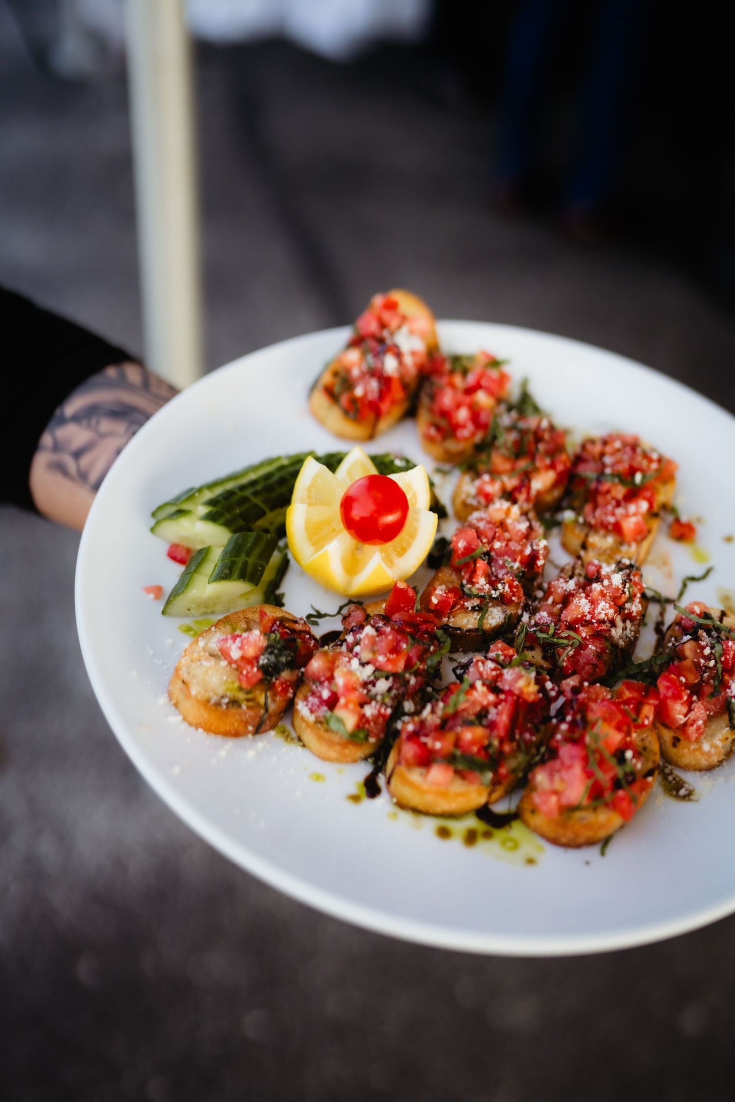 Tattooed hand holding a white plate of bruschetta topped with tomatoes, herbs, lemon, cherry, and cucumber slices.
