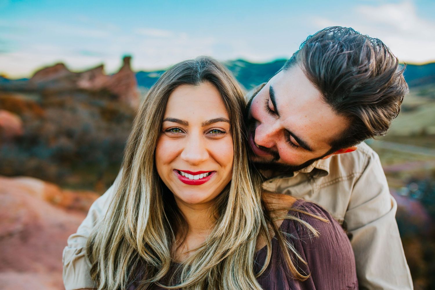 A smiling couple poses outdoors with rocky desert landscape, woman with long blonde hair and red lips, man embracing her from behind.